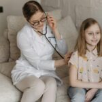 Doctor using a stethoscope for a child's check-up on a sofa during a home visit.